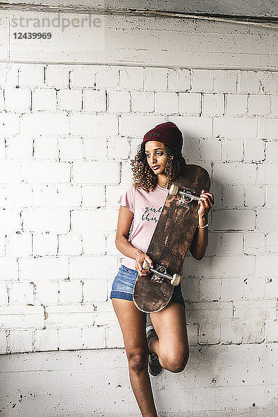 Portrait of a young woman with skateboard  leaning against wall