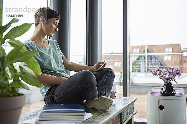 Smiling woman sitting at the window at home using tablet
