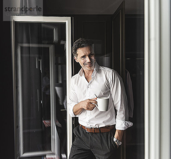 Smiling businessman with cup of coffee standing at the window at home