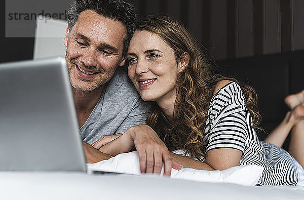 Happy couple lying on bed at home looking at laptop