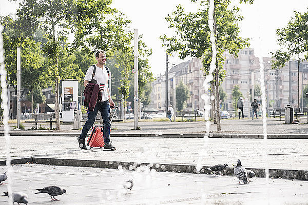 Man with rolling suitcase and takeaway coffee walking at fountain in the city