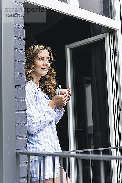 Smiling woman in pyjama at home with cup of coffee looking out of balcony door