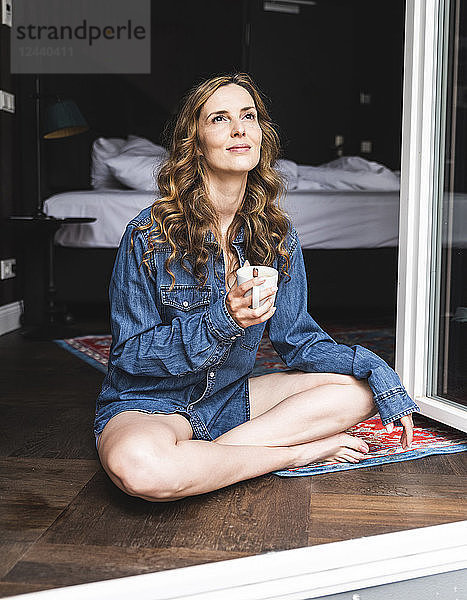 Smiling woman in denim shirt at home with cup of coffee sitting at French window