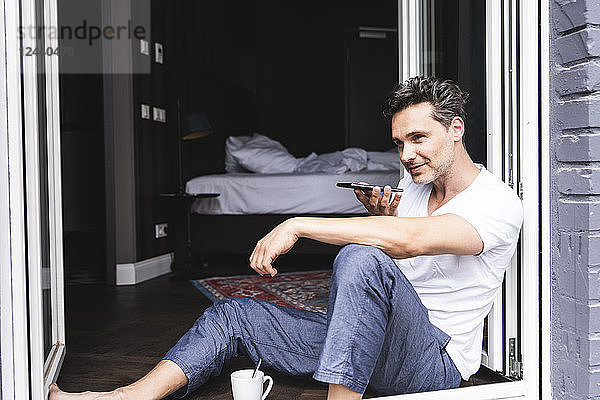 Man in pyjama at home sitting at French window using smartphone