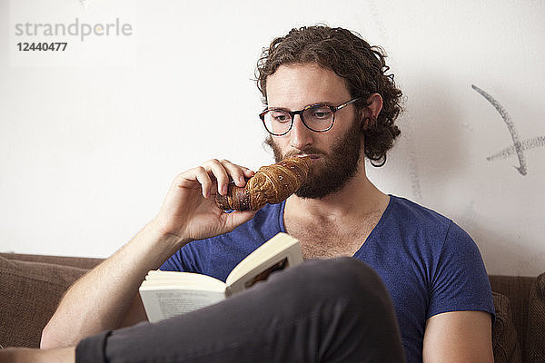Portrait of young man eating croissant in a coffee shop while reading a book