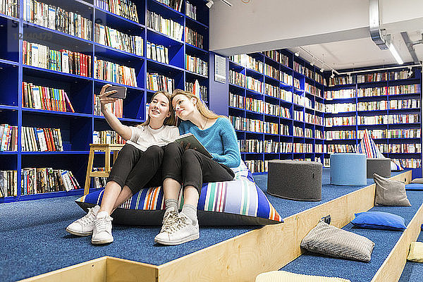 Two teenage girls sitting on beanbag in a public library taking selfie with smartphone