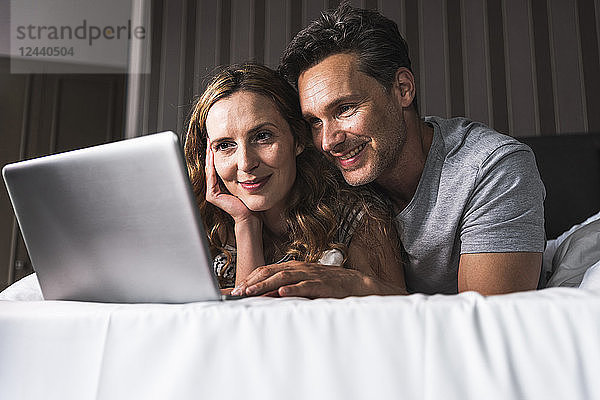 Smiling couple lying on bed at home looking at laptop