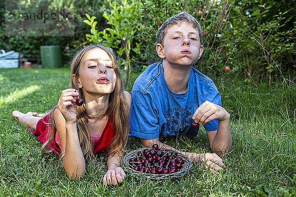 Portrait of brother and sister lying on a meadow spitting cherry pits