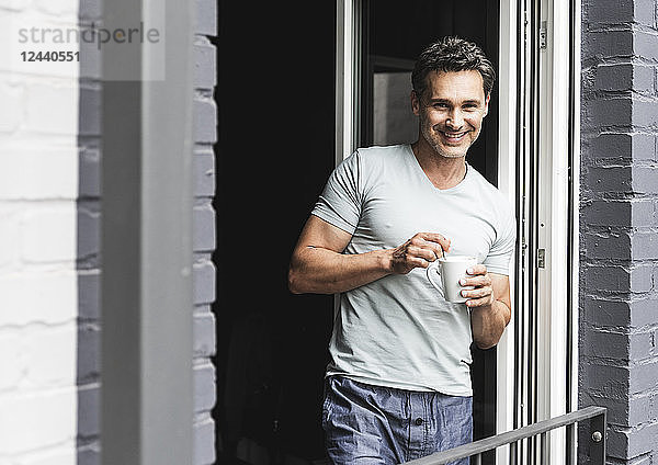Portrait of smiling man in pyjama at home with cup of coffee at balcony door