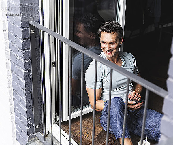 Smiling man in pyjama at home with cell phone looking out of balcony door