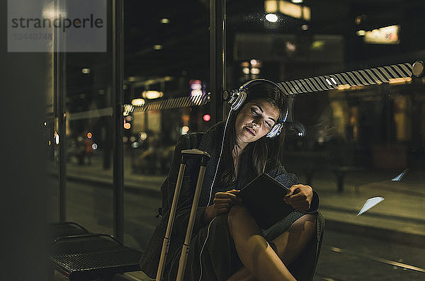 Portrait of young woman with headphones and tablet waiting at station by night