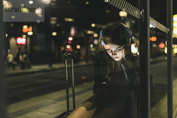 Young woman with headphones waiting at the station by night using tablet