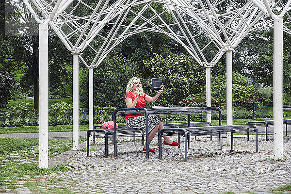 Smiling pregnant woman sitting on a park bench taking a selfie with a tablet