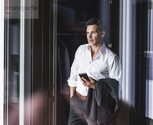Businessman with cell phone standing at wardrobe at home