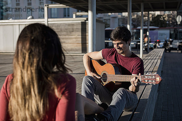 Smiling man sitting on a bench playing guitar for girlfriend