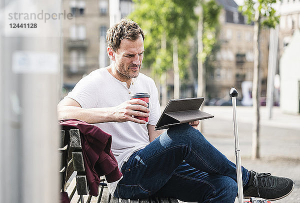 Man with rolling suitcase and takeaway coffee sitting on bench using tablet