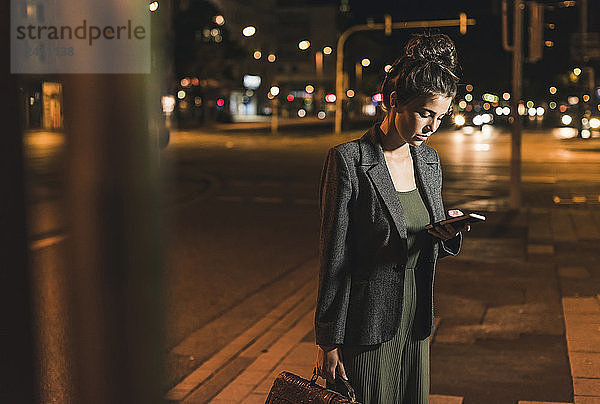Young businesswoman with leather bag looking at cell phone by night