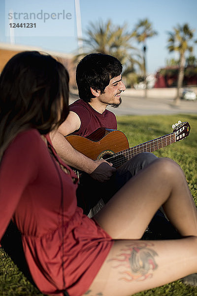 Spain  Barcelona  smiling young man playing guitar for girlfriend sitting on a meadow