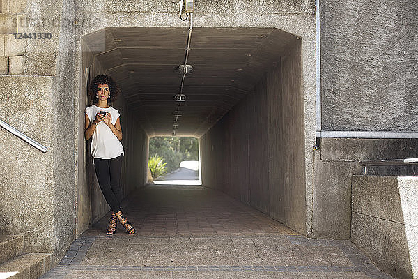 Young woman with smartphone leaning in underpass