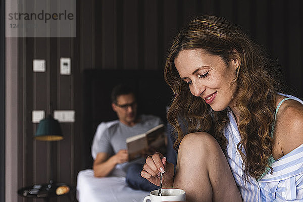Smiling woman with cup of coffee in bedroom with man in background