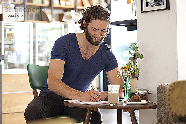 Smiling young man sitting in a coffee shop writing letter