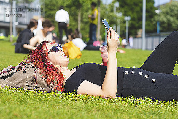 Young woman with smartphone and smoothie relaxing on a meadow
