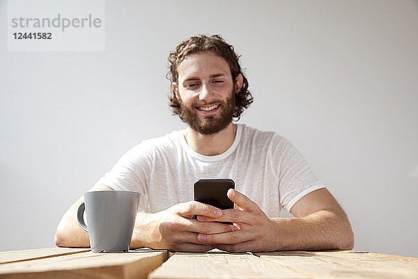 Portrait of smiling man sitting with coffee mug on balcony using cell phone