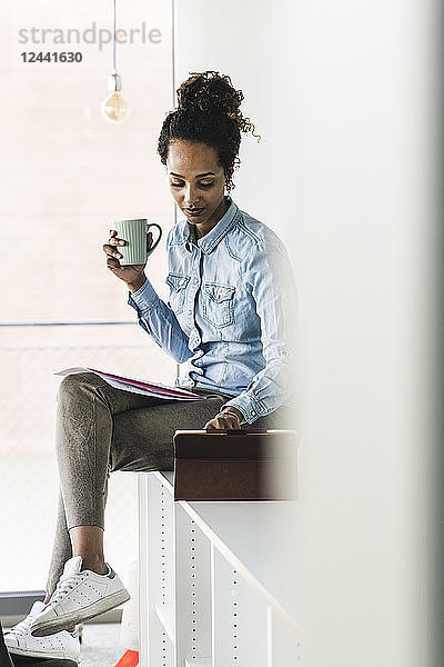 Young businesswoman sitting on shelf  using digital tablet  drinking coffee
