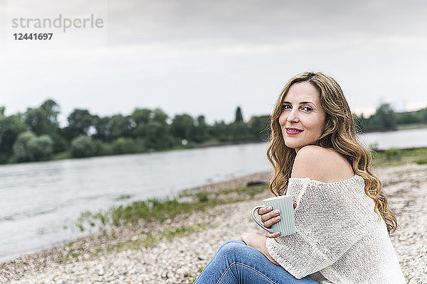 Smiling woman with cup of coffee sitting at the riverside