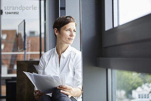 Businesswoman sitting in office with documents looking out of window