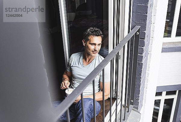 Man in pyjama at home with cup of coffee looking out of balcony door