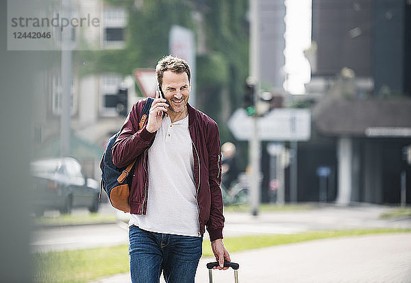 Smiling man with rolling suitcase on cell phone in the city