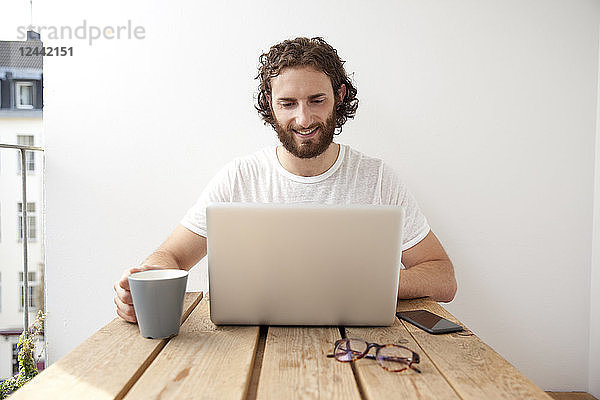 Portrait of smiling man sitting with coffee mug on balcony using laptop