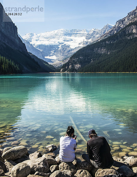 Zwei Personen  die durch den Blick auf ihre Smartphones abgelenkt sind  während sie am Ufer des Lake Louise sitzen; Alberta Kanada