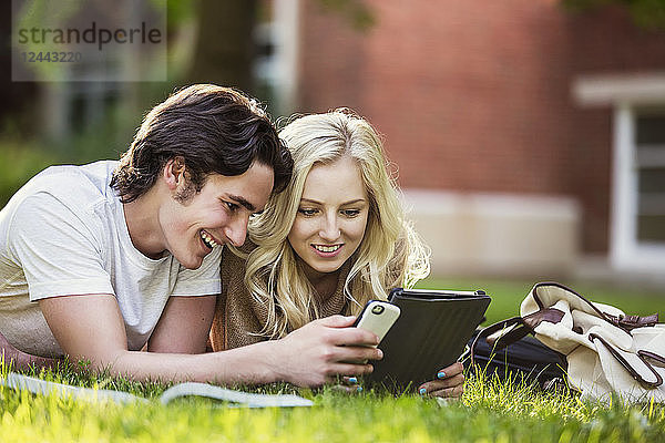 Ein junges Paar studiert im Freien auf dem Rasen des Universitätscampus mit einem Lehrbuch und einem Tablet und überprüft die sozialen Medien auf einem Smartphone  Edmonton  Alberta  Kanada