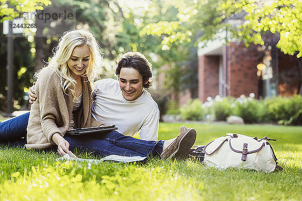 Ein junges Paar  das im Freien auf dem Rasen des Universitätsgeländes studiert und mit einem Tablet in einem Lehrbuch blättert  Edmonton  Alberta  Kanada