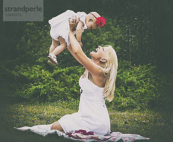 Eine hübsche junge Mutter mit langen blonden Haaren genießt die Zeit mit ihrer süßen kleinen Tochter und wirft sie in die Luft  während sie an einem Sommertag im Gras eines Stadtparks sitzt  Edmonton  Alberta  Kanada