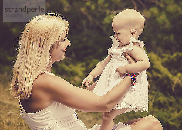 Ein Vintage-Stil Bild von einer schönen jungen Mutter mit langen blonden Haaren genießen die Zeit mit ihrem süßen Baby Tochter sitzen auf dem Rasen in einem Stadtpark an einem Sommertag  Edmonton  Alberta  Kanada