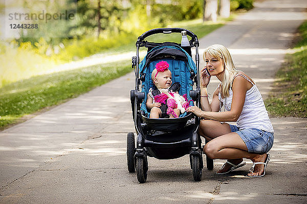 Eine hübsche junge Mutter geht mit ihrer kleinen Tochter im Kinderwagen in einem Park mit einem See an einem warmen sonnigen Tag spazieren und hält an  um einen Anruf mit ihrem Handy entgegenzunehmen  Edmonton  Alberta  Kanada