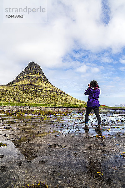 Eine Wanderin hält bei Ebbe am Strand an  um ein Foto mit einer Spiegelreflexkamera vor dem Berg Kirkjufell auf der Halbinsel Snaefellsnes  Westisland  Island  zu machen