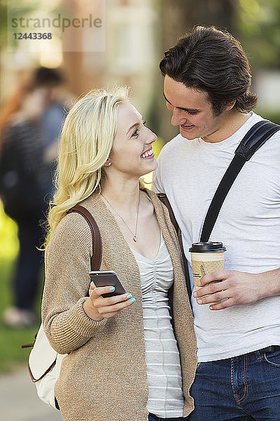 Ein junges Paar  das sich in die Augen schaut und soziale Medien auf einem Smartphone abruft  während es durch einen Universitätscampus geht  Edmonton  Alberta  Kanada