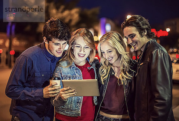 Eine Gruppe von vier Freunden kauert auf einem Bürgersteig zusammen und schaut auf ein Tablet  während das Leuchten des Bildschirms ihre Gesichter erhellt  Edmonton  Alberta  Kanada