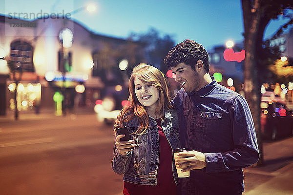 Ein junges Paar  das in der Abenddämmerung auf einem Bürgersteig in einer beliebten Gegend spazieren geht und dabei auf sein Smartphone schaut  Edmonton  Alberta  Kanada