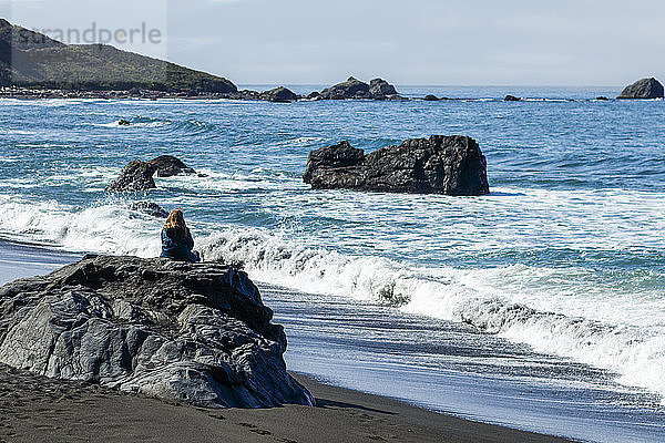 Eine Frau sitzt auf einem Felsen am Strand und beobachtet die Wellen  die an der felsigen kalifornischen Küste an Land kommen  Crescent City  Kalifornien  Vereinigte Staaten von Amerika