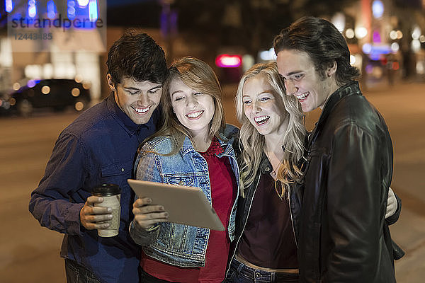 Eine Gruppe von vier Freunden kauert auf einem Bürgersteig zusammen und schaut auf ein Tablet  während das Leuchten des Bildschirms ihre Gesichter erhellt  Edmonton  Alberta  Kanada