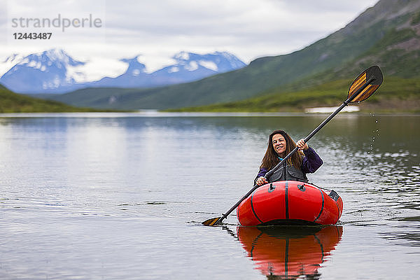 Eine Frau paddelt mit einem Packraft über den Landmark Gap Lake  abseits des Denali Highway  mit der Alaska Range in der Ferne  Alaska  Vereinigte Staaten von Amerika
