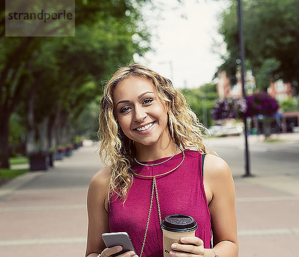 Eine junge Frau blickt in die Kamera  während sie mit einem Kaffee in der Nähe des Universitätsgeländes eine Straße entlanggeht und eine SMS auf ihrem Smartphone schreibt  Edmonton  Alberta  Kanada