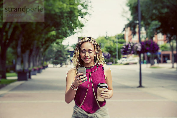 Eine schöne junge Frau  die eine Straße in der Nähe eines Universitätscampus entlangläuft und eine SMS auf ihrem Smartphone schreibt  Edmonton  Alberta  Kanada