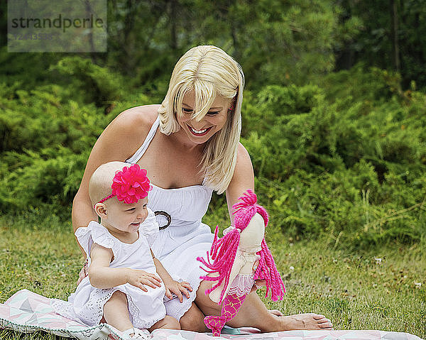 Eine hübsche junge Mutter genießt die Zeit mit ihrer süßen kleinen Tochter  die auf einer Decke sitzt und mit einer Puppe in einem Stadtpark an einem Sommertag spielt  Edmonton  Alberta  Kanada.