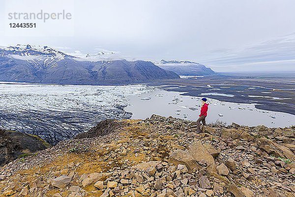 Eine Wanderin in warmer Kleidung für die Elemente steht am Rand einer Bergklippe mit Blick auf den Gletschersee und das Tal im Vatnajokull-Nationalpark  Island